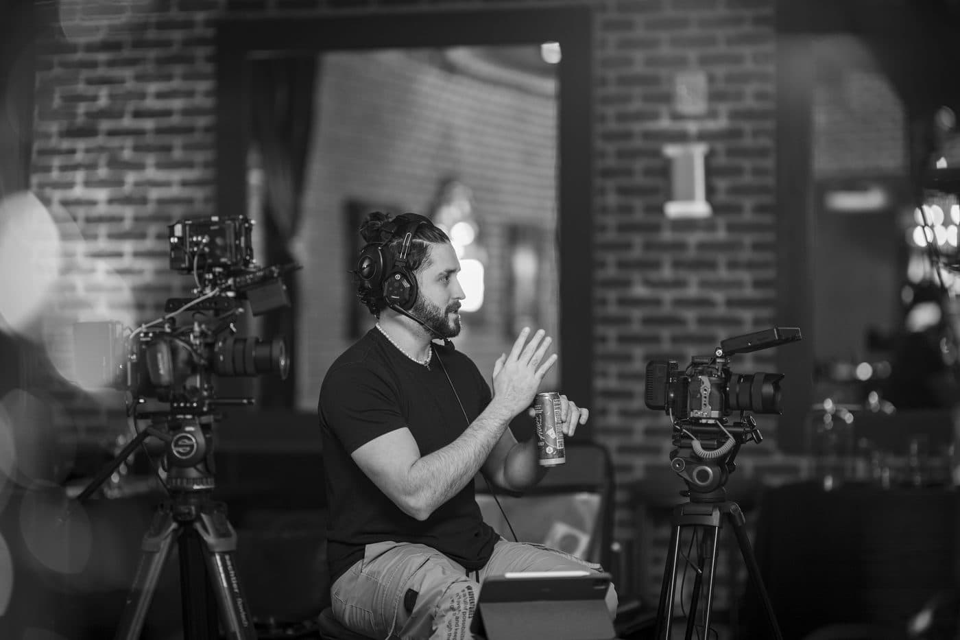 Black and white photo of a Hyped Visuals director seated between two cinema cameras on tripods, gesturing as he talks