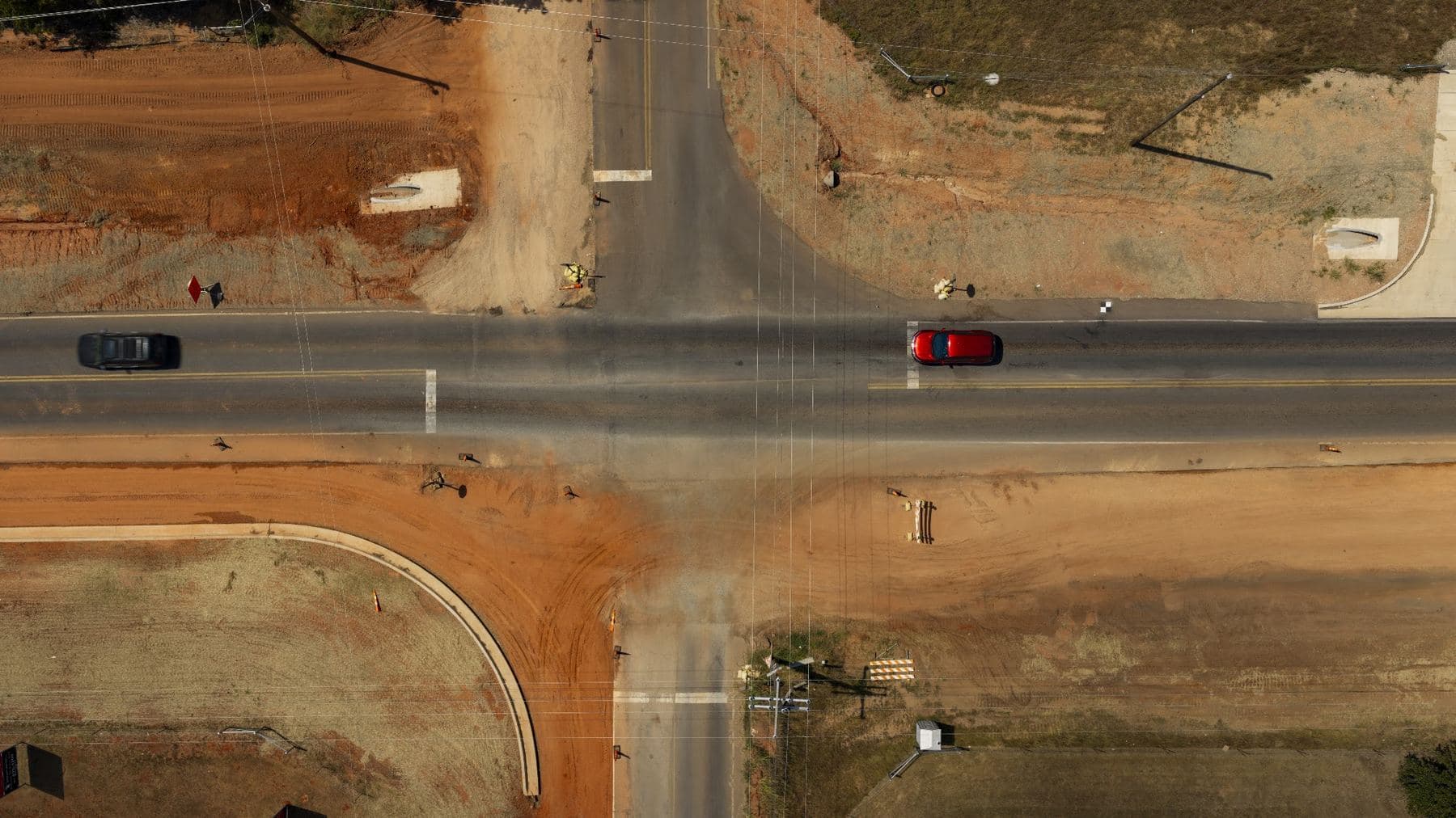 Straight down aerial view of a roadway intersection under construction, red soil graded around new curbs