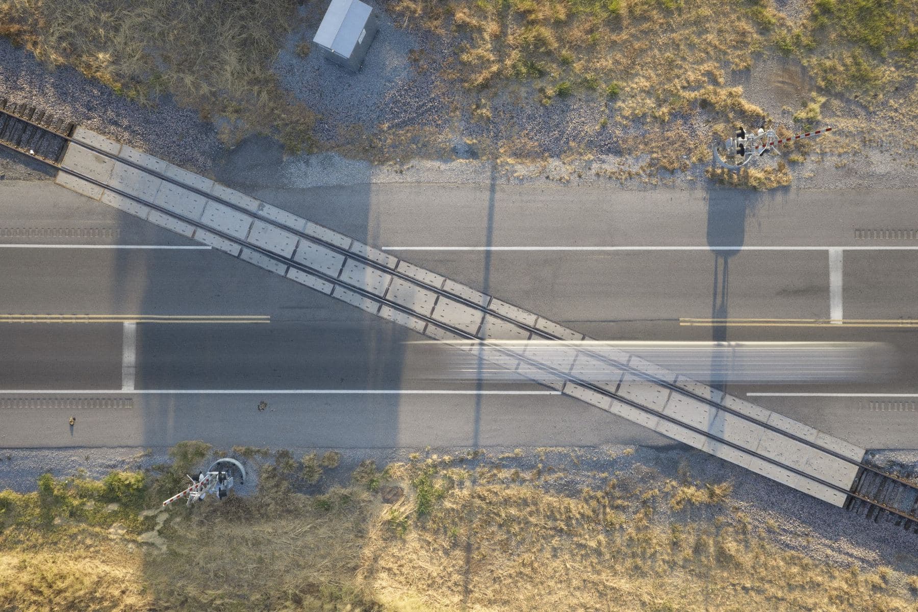 Overhead view of a narrow bridge structure crossing a highway at an angle, its shadow falling across the lanes