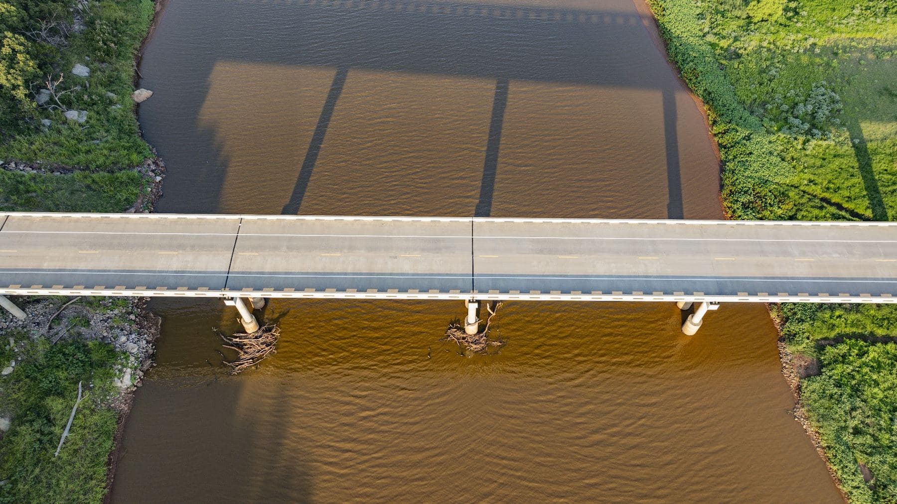 Straight down aerial view of a highway bridge crossing a muddy river, its shadow and piers visible on the water