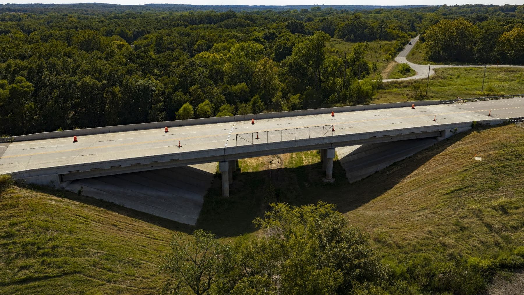 Aerial view of a concrete highway bridge spanning a wooded drainage, with a rural road curving away behind it