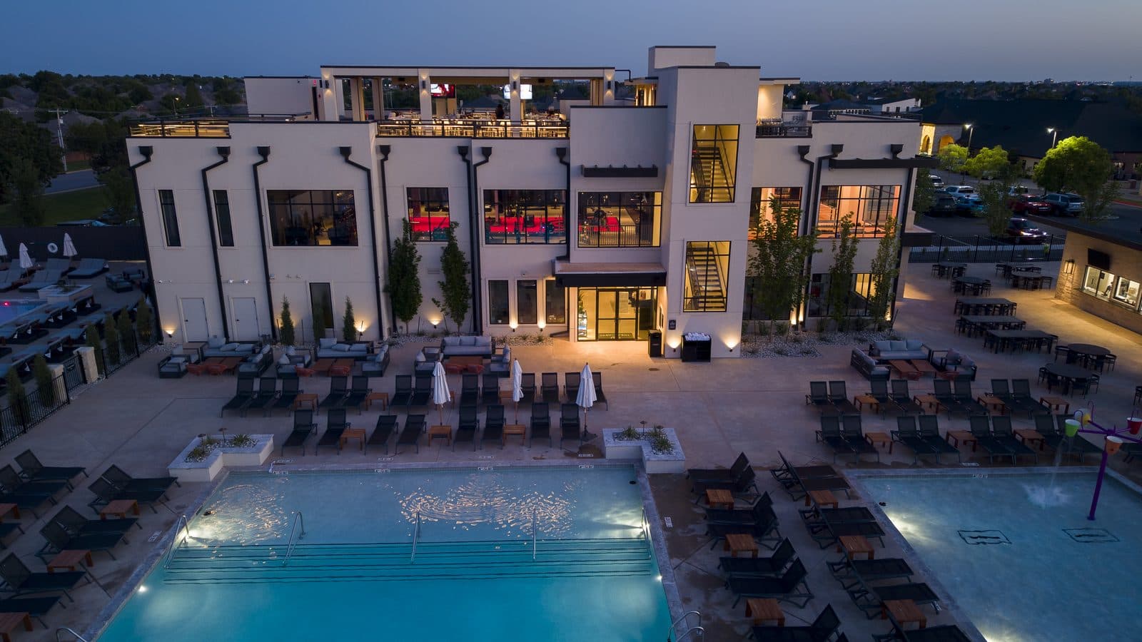 Aerial view at dusk of a lit three story clubhouse with a rooftop deck above a pool and splash pad at Rose Creek