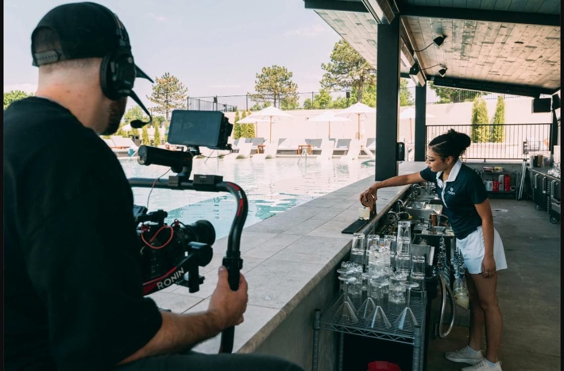 A Hyped Visuals camera operator in headphones films with a gimbal rig as a Rose Creek staff member works the poolside bar
