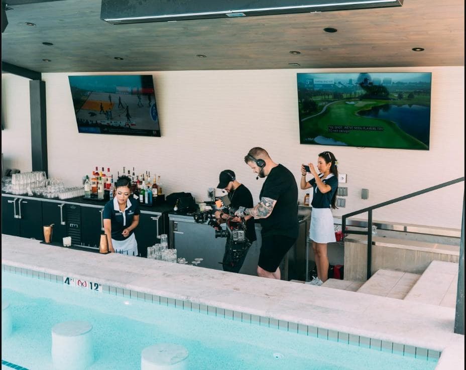 Two Hyped Visuals crew members film with a gimbal camera rig behind Rose Creek's swim up bar while two staff members work nearby