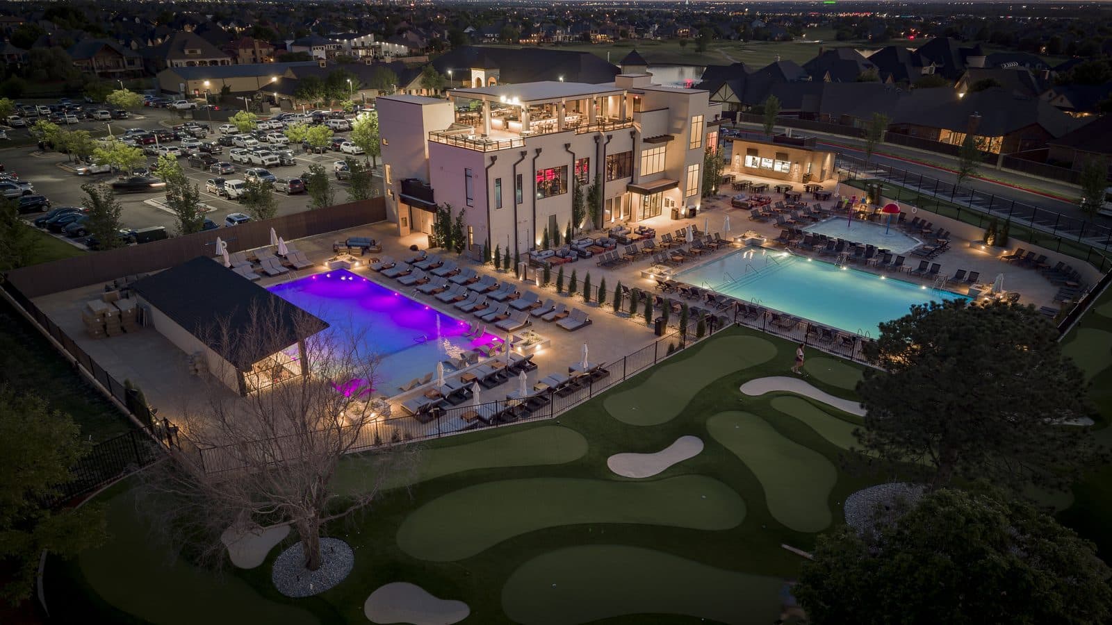 Aerial view at dusk of Rose Creek Country Club's lit clubhouse, two pools and putting green in Edmond, Oklahoma